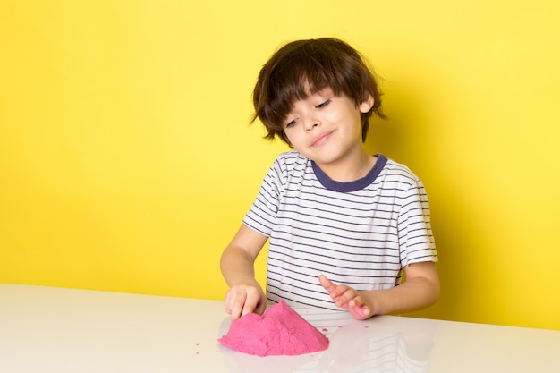 Una vista frontal lindo niño adorable en camiseta a rayas jugando con arena cinética colorida