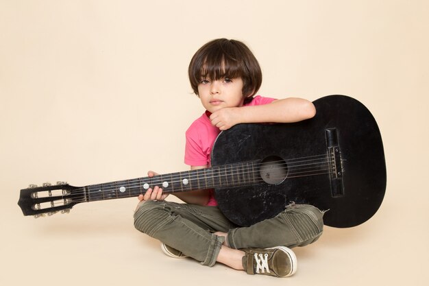 Una vista frontal lindo niño deprimido en camiseta rosa jeans caqui tocando la guitarra negra