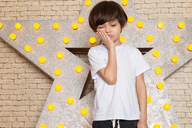 Foto gratuita una vista frontal lindo niño pequeño en camiseta blanca jeans oscuros en la estrella diseñada soporte amarillo y fondo claro