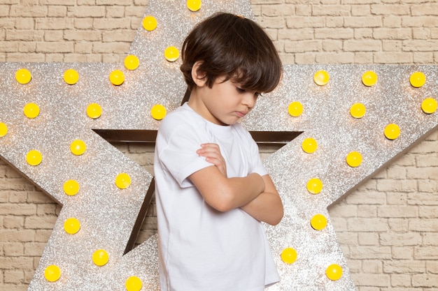 Una vista frontal lindo niño pequeño en camiseta blanca jeans oscuros en la estrella diseñada soporte amarillo y fondo claro