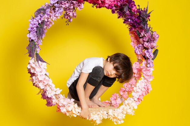 Una vista frontal lindo niño pequeño en camiseta blanca sentado en las flores hechas pararse en el piso amarillo