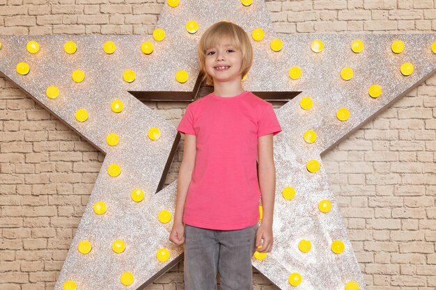 Una vista frontal lindo niño pequeño en camiseta rosa jeans grises sonriendo en la estrella diseñada soporte amarillo y fondo claro