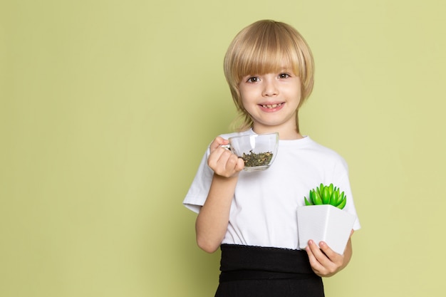 Una vista frontal lindo niño sonriente en camiseta blanca con especies y pequeña planta verde en el escritorio de color piedra