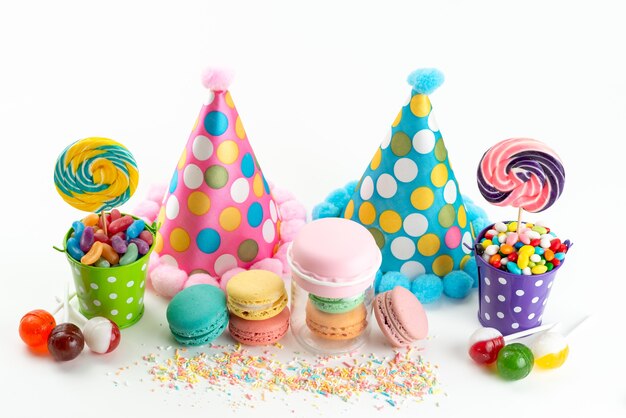 Una vista frontal macarons franceses coloridos caramelos y piruletas junto con divertidos gorros de cumpleaños en blanco, celebración de color dulce de azúcar