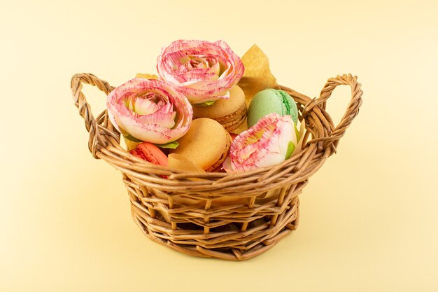 Una vista frontal de macarons franceses con flores dentro de la canasta en el escritorio amarillo bizcocho de azúcar dulce