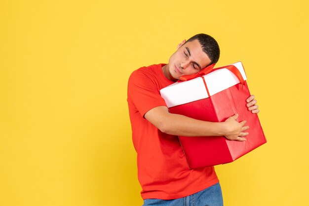 Vista frontal macho joven abrazando el regalo de Navidad sobre fondo amarillo