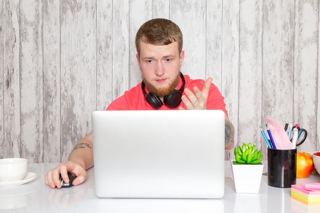 Una vista frontal macho joven en camisa rosa en auriculares negros con mesa gris portátil con bolígrafos planta café espacio gris