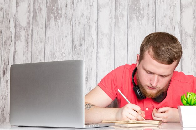 Una vista frontal macho joven en camisa rosa en auriculares negros usando laptop gris escribiendo notas mesa con bolígrafos planta de café espacio gris