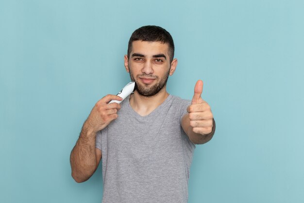 Vista frontal macho joven en camiseta gris afeitándose la barba en espuma masculina de barba de afeitado azul hielo