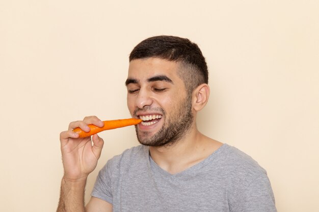 Vista frontal macho joven en camiseta gris comiendo zanahoria en beige ¡