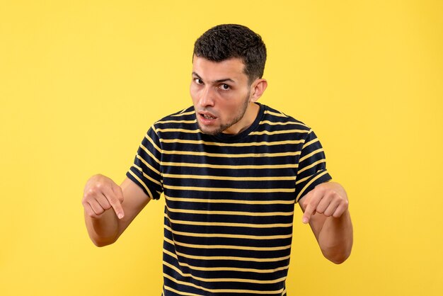 Vista frontal macho joven en camiseta a rayas blanco y negro apuntando al piso fondo amarillo aislado