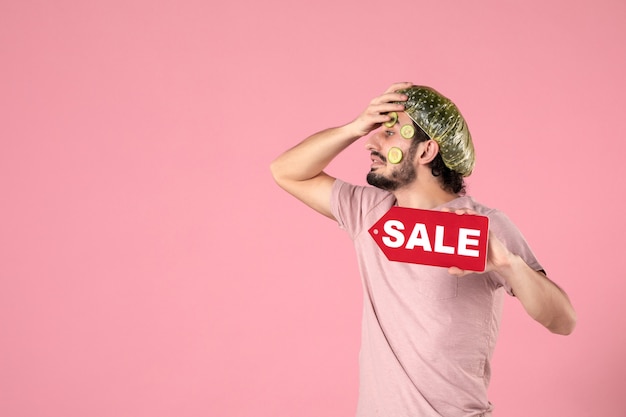 Vista frontal macho joven con máscara en su rostro sosteniendo placa de venta sobre fondo de color rosa