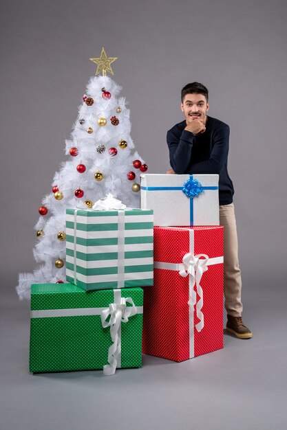 Vista frontal macho joven con regalos y árbol de Navidad en gris