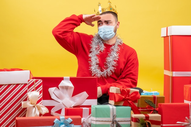 Vista frontal macho joven sentado alrededor de regalos de Navidad en máscara sobre fondo amarillo