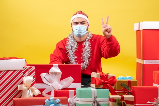 Vista frontal macho joven sentado alrededor de regalos de Navidad en máscara sobre fondo amarillo