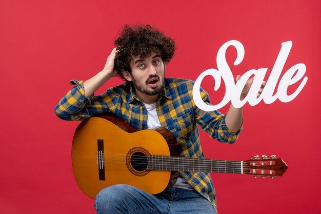 Vista frontal macho joven sentado con guitarra en venta de pared roja tocar música de concierto color músico en vivo