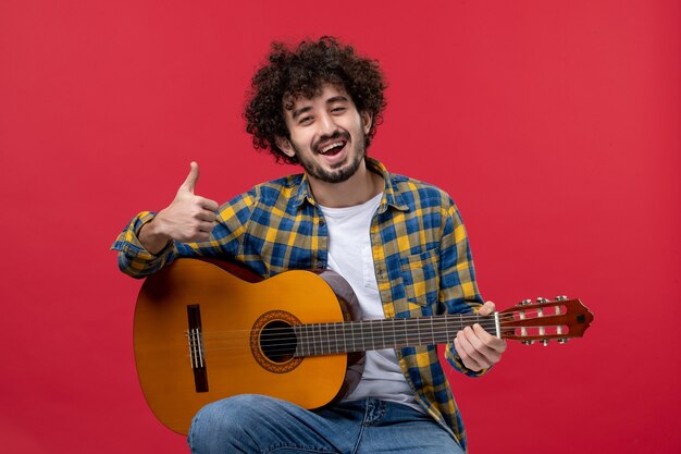 Vista frontal macho joven sentado y tocando la guitarra en la pared roja concierto banda de color música tocar músico aplauso