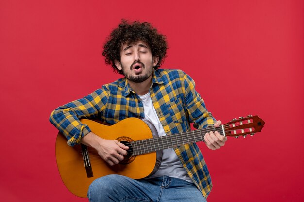 Vista frontal macho joven sentado y tocando la guitarra en la pared roja concierto en vivo músico de color banda de aplausos reproducir música