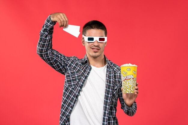 Vista frontal macho joven sosteniendo boletos de palomitas de maíz en -d gafas de sol en la pared de color rojo claro cine masculino cine película