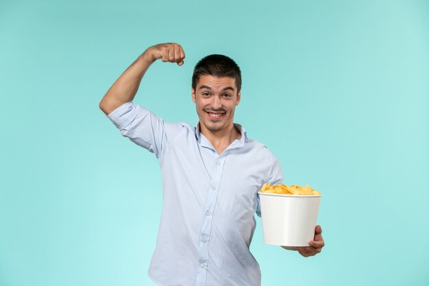 Vista frontal macho joven sosteniendo la canasta con papas fritas y flexionando sobre la superficie azul