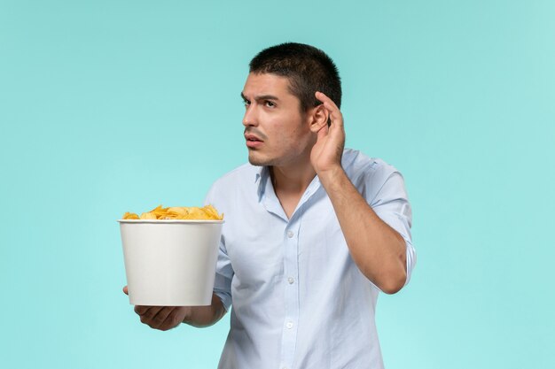 Vista frontal macho joven sosteniendo la canasta con papas fritas y tratando de escuchar sobre una superficie azul