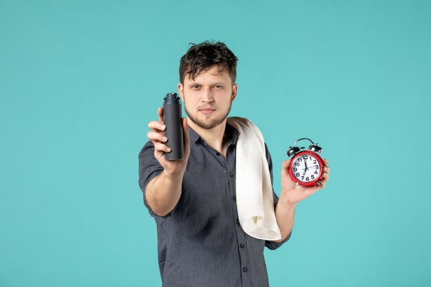 Vista frontal macho joven sosteniendo espuma para afeitarse y reloj sobre fondo azul.