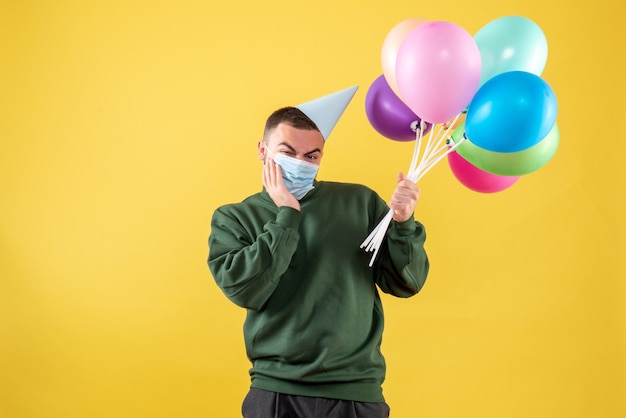 Vista frontal macho joven sosteniendo globos de colores en máscara sobre fondo amarillo