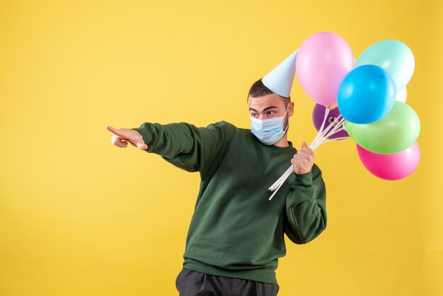 Vista frontal macho joven sosteniendo globos de colores en máscara sobre fondo amarillo