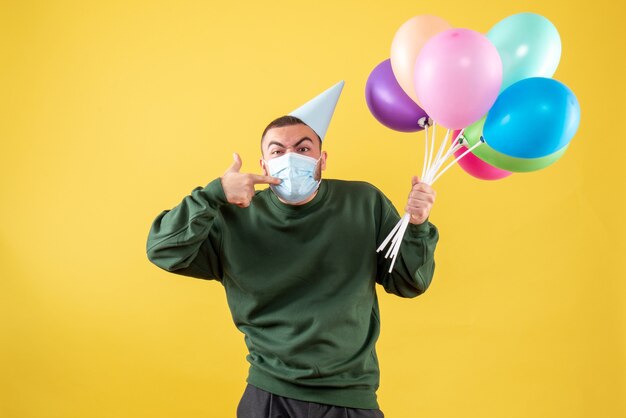 Vista frontal macho joven sosteniendo globos de colores en máscara sobre fondo amarillo