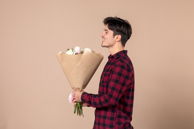 Vista frontal macho joven sosteniendo hermosas flores como presente del día de la mujer de marzo en la pared marrón