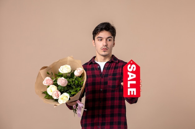 Vista frontal macho joven sosteniendo hermosas flores para mujer y placa de venta en pared marrón