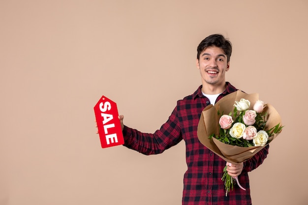 Vista frontal macho joven sosteniendo hermosas flores y placa de venta en pared marrón