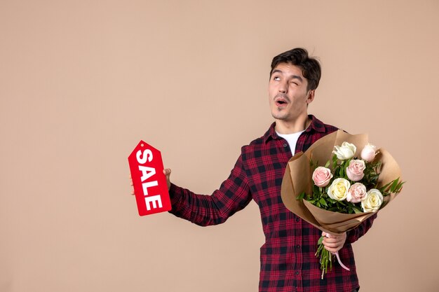 Vista frontal macho joven sosteniendo hermosas flores y placa de venta en pared marrón