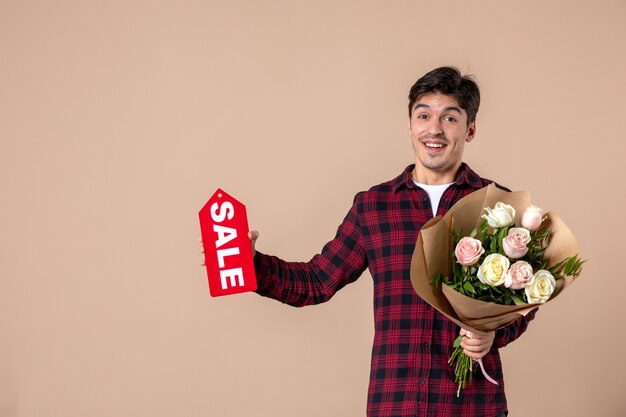 Vista frontal macho joven sosteniendo hermosas flores y placa de venta en pared marrón