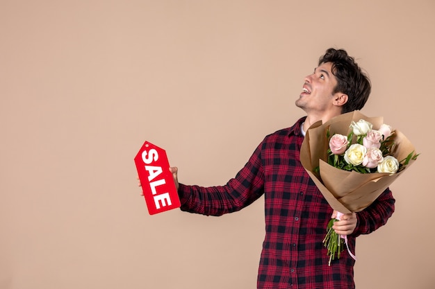 Vista frontal macho joven sosteniendo hermosas flores y placa de venta en pared marrón