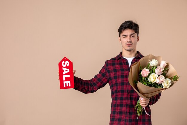 Vista frontal macho joven sosteniendo hermosas flores y placa de venta en pared marrón