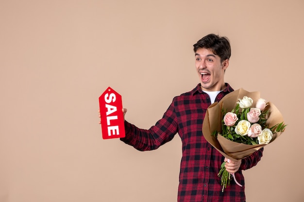 Vista frontal macho joven sosteniendo hermosas flores y placa de venta en pared marrón