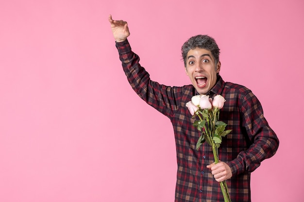 Vista frontal macho joven sosteniendo hermosas rosas rosadas en la pared rosada