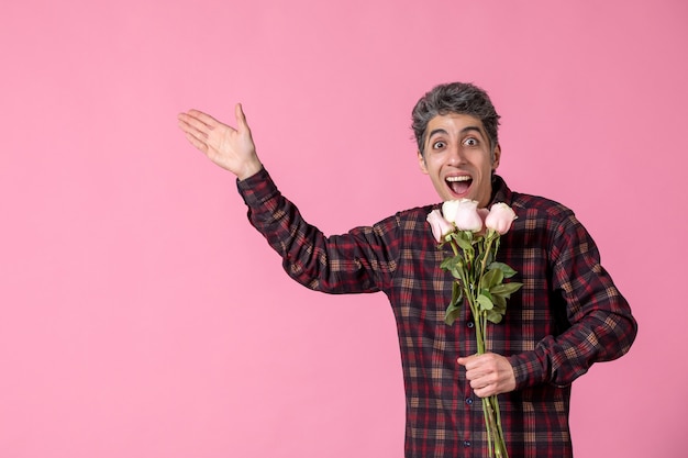 Vista frontal macho joven sosteniendo hermosas rosas rosadas en la pared rosada