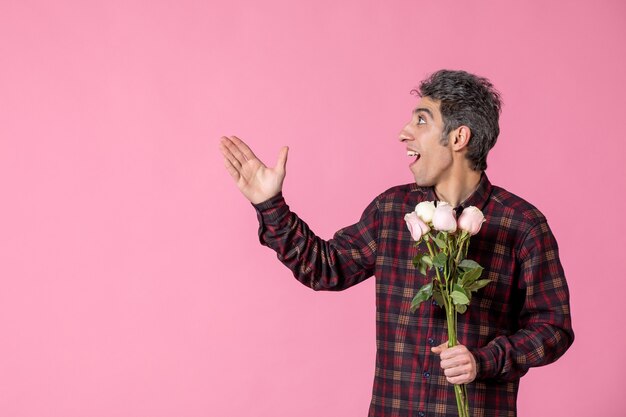 Vista frontal macho joven sosteniendo hermosas rosas rosadas en la pared rosada