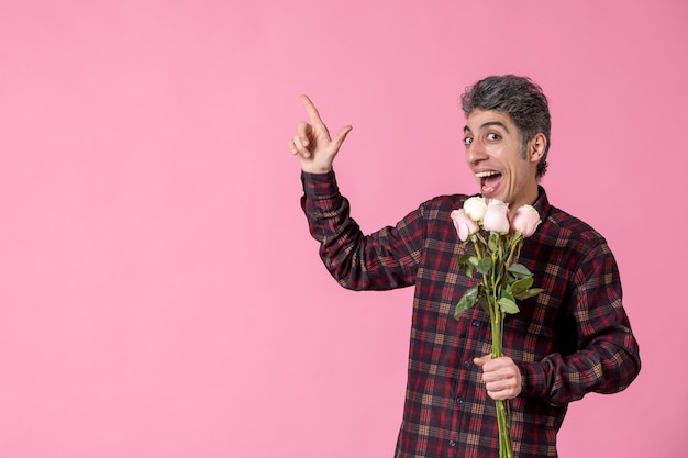 Vista frontal macho joven sosteniendo hermosas rosas rosadas en la pared rosada