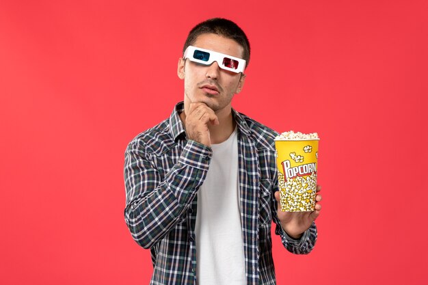 Vista frontal macho joven sosteniendo el paquete de palomitas de maíz en -d gafas de sol pensando en la pared de color rojo claro cine cine cine película