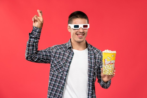 Vista frontal macho joven sosteniendo el paquete de palomitas de maíz y con gafas de sol -d en la pared de color rojo claro cine película de cine