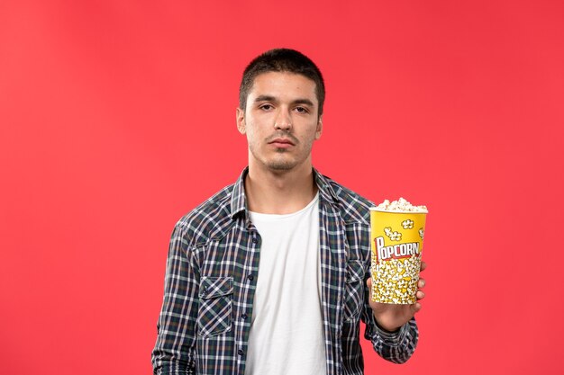 Vista frontal macho joven sosteniendo el paquete de palomitas de maíz en la pared roja cine cine película de cine