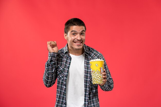 Vista frontal macho joven sosteniendo el paquete de palomitas de maíz y regocijándose en la pared de color rojo claro cine cine película masculina película
