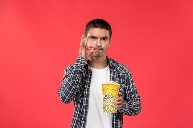 Vista frontal macho joven sosteniendo el paquete de palomitas de maíz y sonriendo en el escritorio rojo claro cine películas teatro película chico masculino