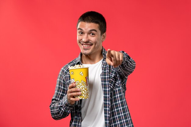 Vista frontal macho joven sosteniendo el paquete de palomitas de maíz y sonriendo en la pared de color rojo claro cine películas teatro película chico masculino