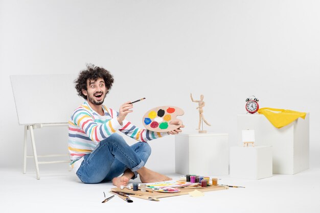 Vista frontal del macho joven sosteniendo pinturas y borla para dibujar en la pared blanca