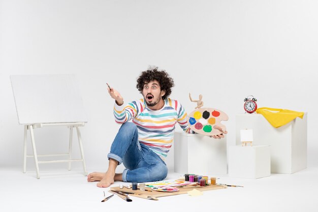 Vista frontal del macho joven sosteniendo pinturas y borla para dibujar en la pared blanca