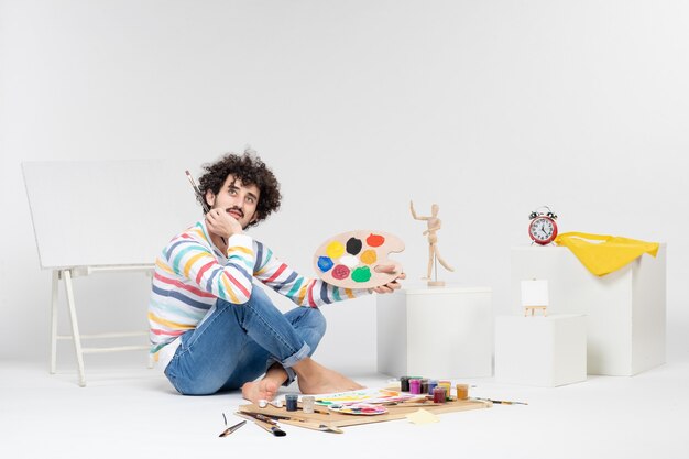 Vista frontal del macho joven sosteniendo pinturas y borla para dibujar en la pared blanca